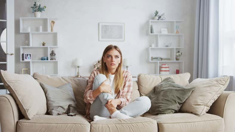 Sad woman is sitting on comfortable sofa with crossed legs and hugging her knees, she is crying while watching a drama movie or tv show in her living room