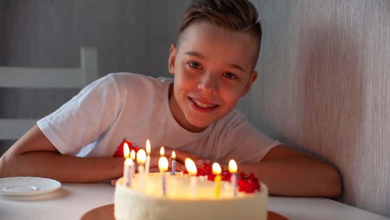 Smiling young boy leaning on a table with a lit birthday cake, enjoying a joyful celebration in a cozy setting.