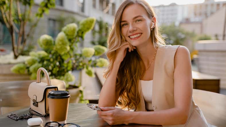 attractive young blond woman sitting in cafe using smart phone drinking coffee in spring summer autumn fashion trend outfit smiling happy wearing beige suit street style, thinking busy and confident