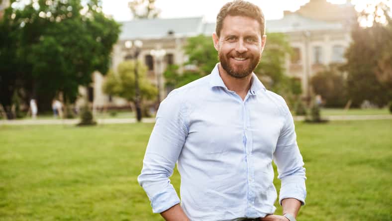 Portrait of european attractive man in white shirt standing with hands in pockets during walk in green park over old building background