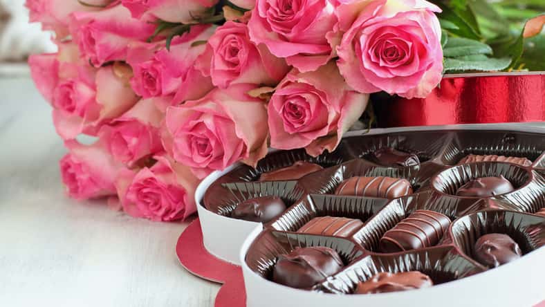 Dozen soft pink rose flowers with a heart shaped box of chocolate candy for Valentine Day over a wood background.