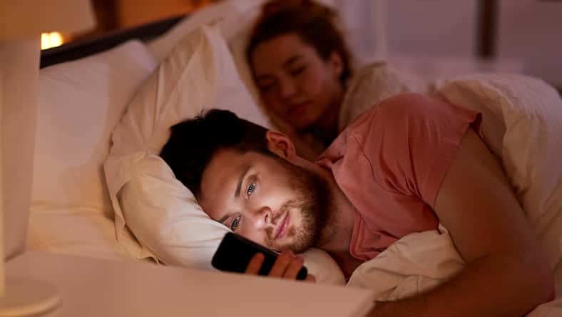 technology, internet addiction and cheat concept - man using smartphone at night while girlfriend is sleeping