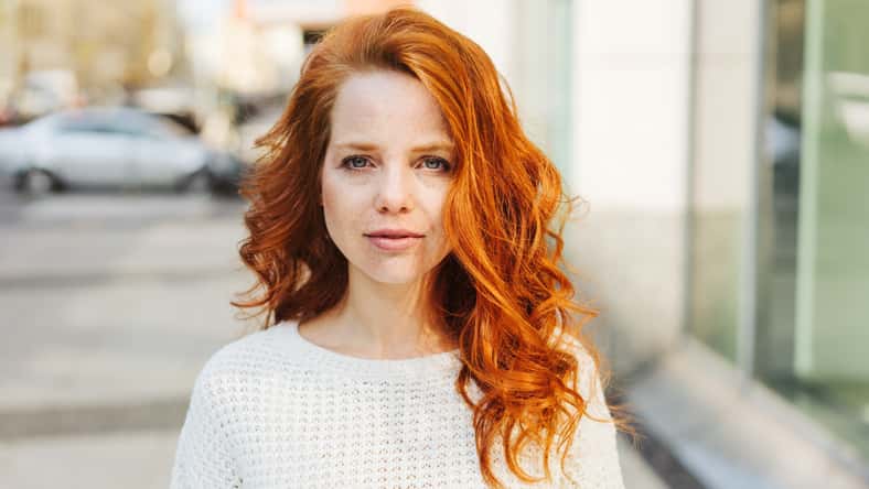 Serious attractive young woman with long red hair looking pensively at the camera as she stands in a city street