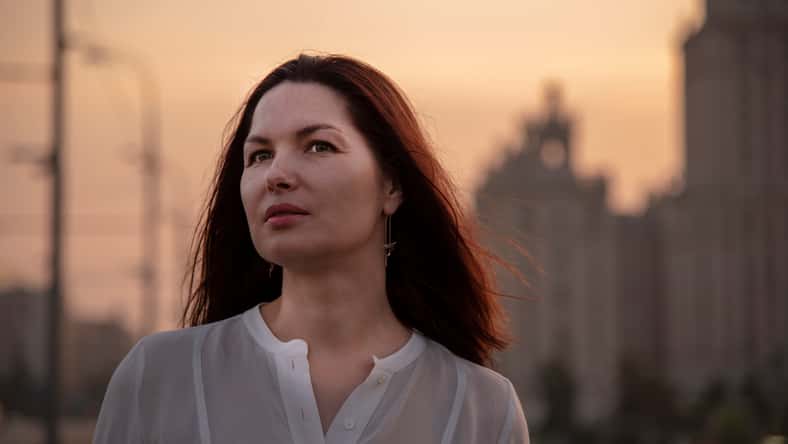 Portrait of a middle-aged woman in profile in a white blouse with her hair down. Against the background of the evening city. A dreamy look into the distance. Houses, sunset.