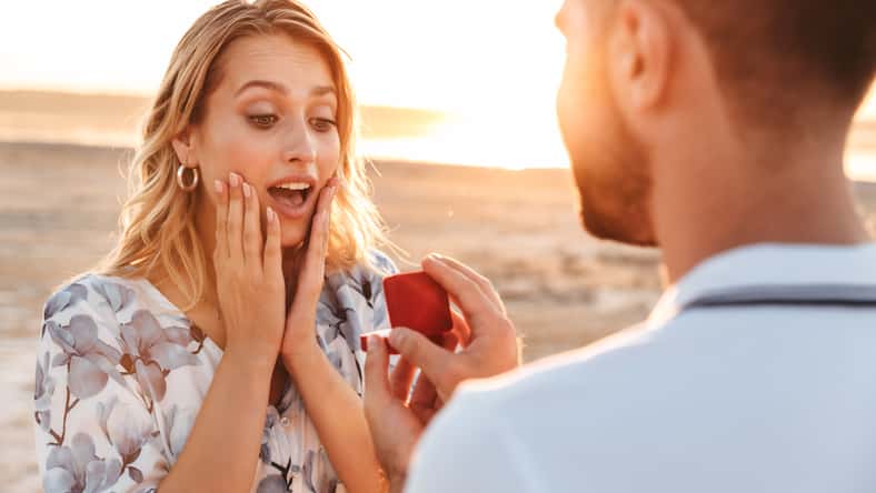 Photo of unshaven man making proposal to his amazed woman with ring in gift box while walking on sunny beach