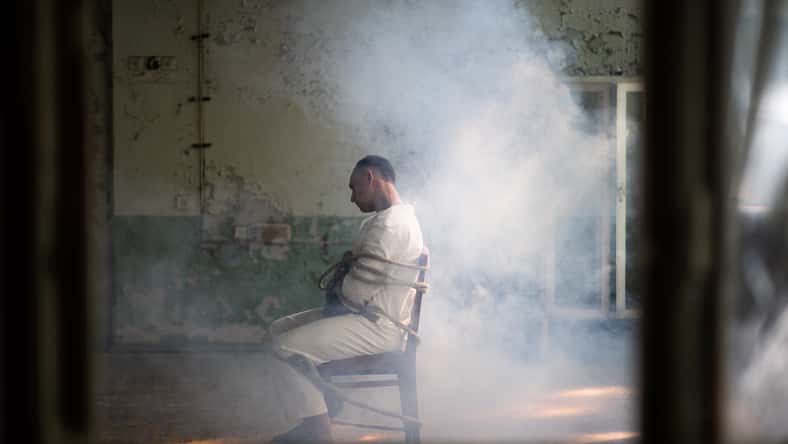 A crazy man in a straitjacket is tied to a chair in an abandoned old clinic.