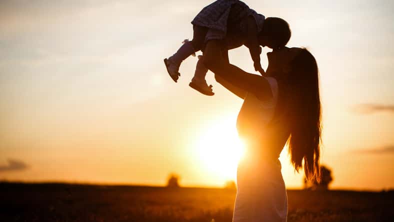 A mother lifts a toddler child in the air above a picturesque sunset sky. A woman and a little girl in a field of lavender flowers
