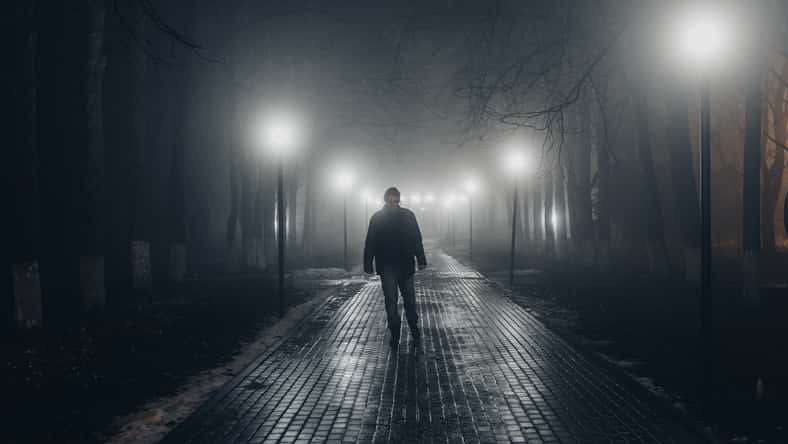 Sad man alone walking along the alley in night foggy park. front view.