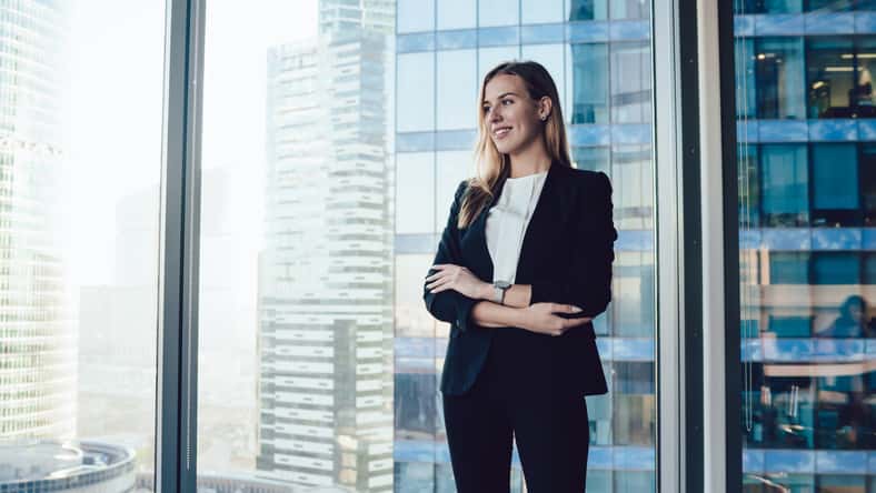 Smiling blonde woman in trendy suit posing in office interior near window with panoramic view on business center, cheerful prosperous female proud ceo in formal outfit standing with crossed hands