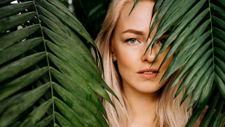 Young woman's face surrounded by tropical leaves. Closeup portrait of magnificent lady with natural nude makeup, perfect skin, blonde hair. Natural cosmetic, wellness, purity, skincare, beauty concept