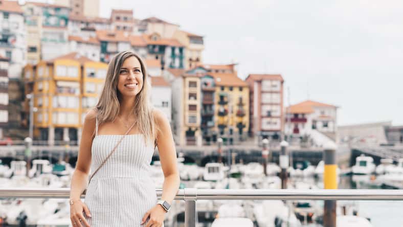 Young woman leaning against a railing, with a port and a small village behind her