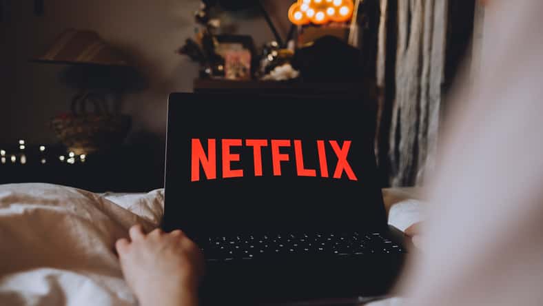 Netflix logo on the laptop screen. Young woman watching netflix channel sitting at home in the evening.