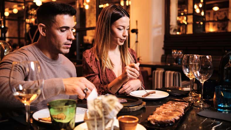 Portrait of young couple enjoying time together and having dinner on a date at fancy restaurant. Couple eating with glasses of wine on table.
