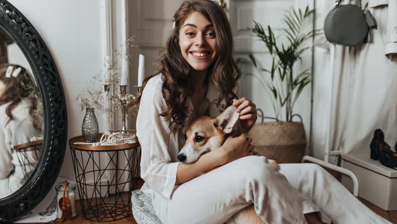 Curly woman in white shirt having fun with dog. Brunette pretty lady in beige blouse and pants plays and hugs corgi while sitting on floor