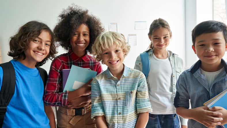 Happy diverse junior school students children group looking at camera standing in classroom. Smiling multiethnic cool kids boys and girls friends posing for group portrait together.