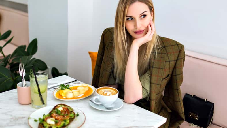Lifestyle indoor portrait of stylish young blonde business woman enjoying her brunch at hipster cafe, asking for waiter and smiling, co working space.