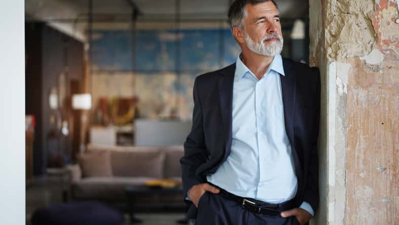 Positive confident mature executive man wearing black suit and blue shirt smiling and looking away while standing in office