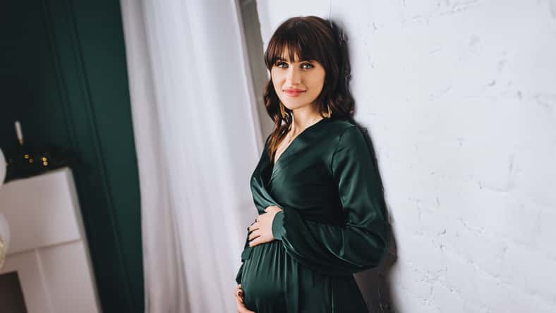 A beautiful pregnant brunette girl in a green dress with a big belly stands near a white wall in the interior.
