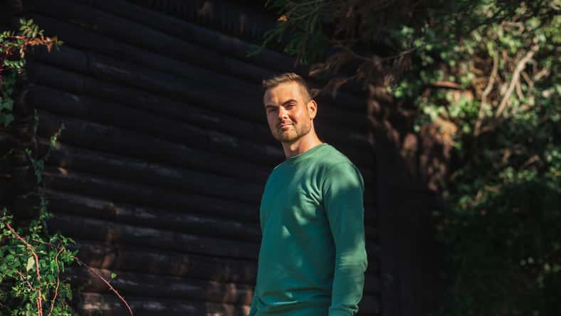 Handsome bearded man in a green sweater posing by log wall and green trees on the sunset. Young eco friendly hipster.