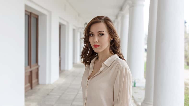 pretty caucasian young woman in beige blouse, red lipstick and wavy hair standing on veranda of cafe. white concrete pillars in background