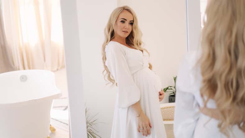 Beautiful portrait of pregnant woman in white dress. Concept of perfect pregnancy. Woman put her hand on belly. Future mother is waiting for baby.