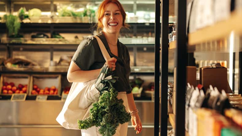 Female customer smiling cheerfully while carrying a bag with fresh organic vegetables in a grocery store. Happy young woman grocery shopping in a trendy supermarket.