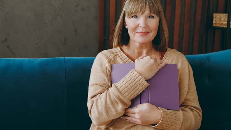 Smiling happy elderly woman 50s years old wearing casual clothes glasses sit on blue sofa read book novel look camera stay at home flat rest relax spend free spare time in living room indoor grey wall