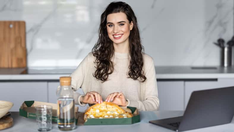 Overworked woman taking a break and eating pizza, working from home on laptop
