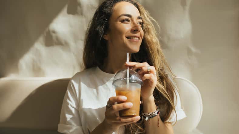 Young beautiful woman with long curly hair drinking iced coffee on beige background.