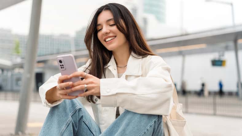 Bright and cheerful young woman with shopper on shoulder waiting public transport at station smiling beautifully while texting on phone.