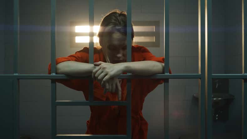 Female prisoner in orange uniform holds hands on metal bars, walks, looks at barred window in jail cell. Woman serves imprisonment term for crime in prison. Depressed inmate in correctional facility.