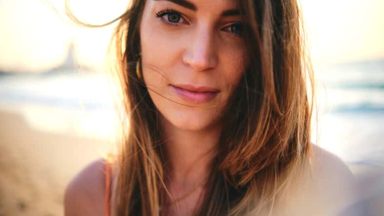 An intimate close-up image of a woman's face with her hair blowing in the wind, on the beach during sunset