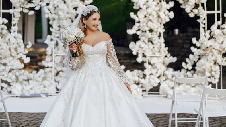 A woman in a white wedding dress is standing in front of a white archway. She is holding a bouquet of flowers and she is posing for a picture. Concept of elegance and celebration