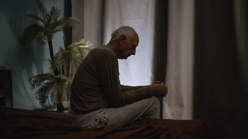 Silhouette depressed senior man 70s sits thoughtful looking down, melancholic male at night on the bed. Copy space