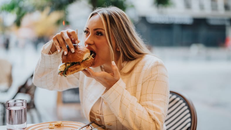 Una mujer está saboreando un sándwich grande mientras cena al aire libre en un acogedor café. El escenario transmite un ambiente relajado y agradable, perfecto para comidas informales y apreciando la deliciosa comida.