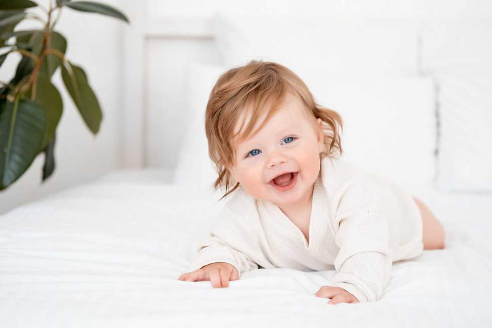 A baby with red hair and blue eyes in a white bodysuit is smiling happily on the bed at home, a healthy happy cute newborn baby boy.