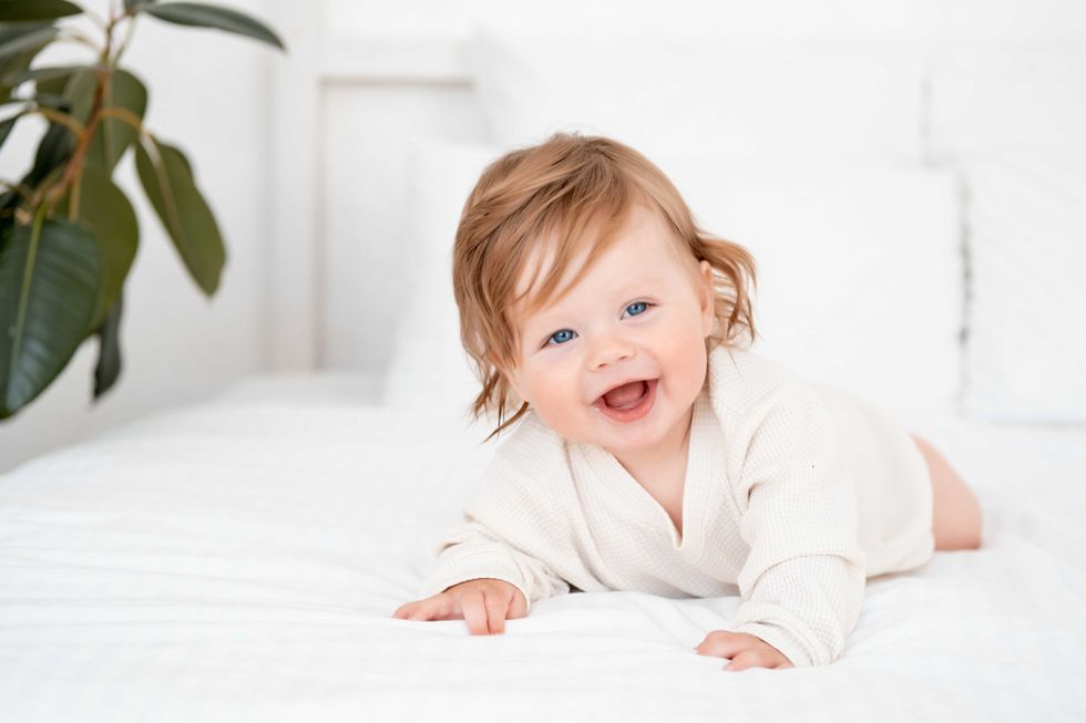 A baby with red hair and blue eyes in a white bodysuit is smiling happily on the bed at home, a healthy happy cute newborn baby boy.