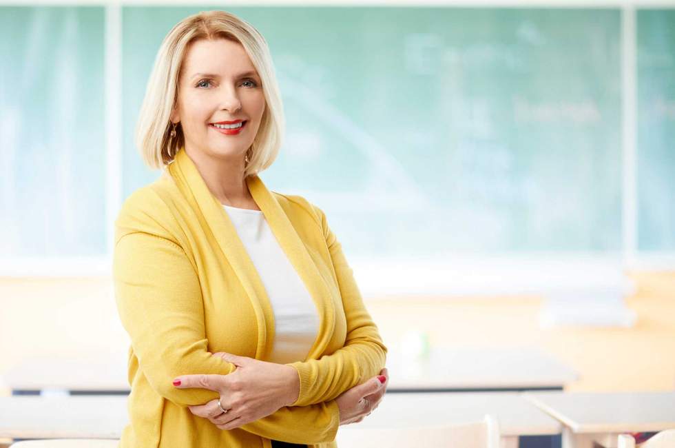 A smiling middle aged female teacher standing at the classroom behind the chalboard.