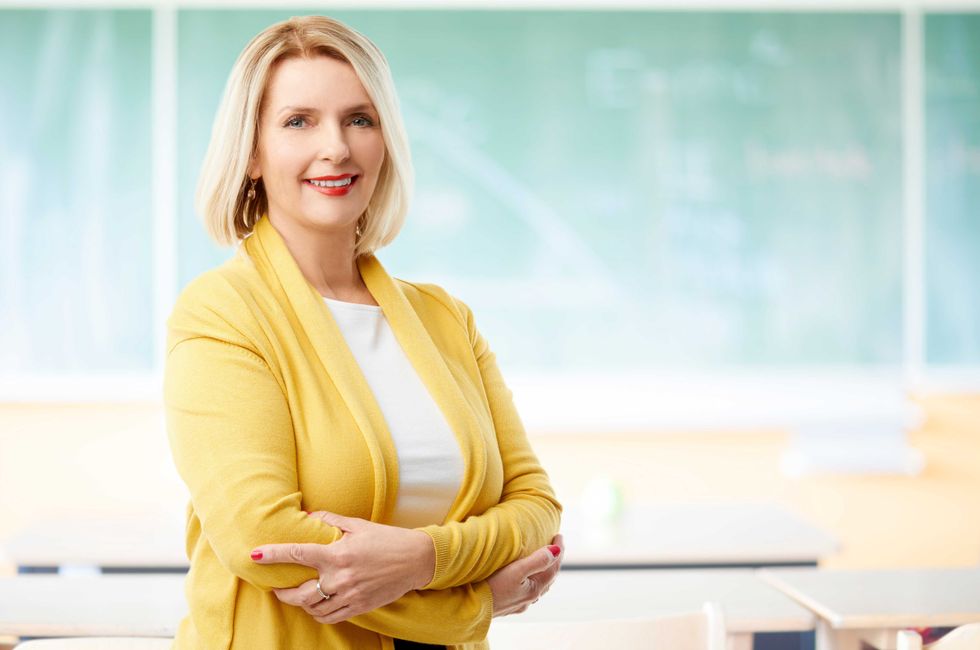 A smiling middle aged female teacher standing at the classroom behind the chalboard.