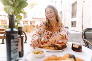 A woman sits at a table in an outdoor cafe. She holds a slice of pizza and smiles. Plates of food and drinks are on the table, and sunlight shines through the trees.
