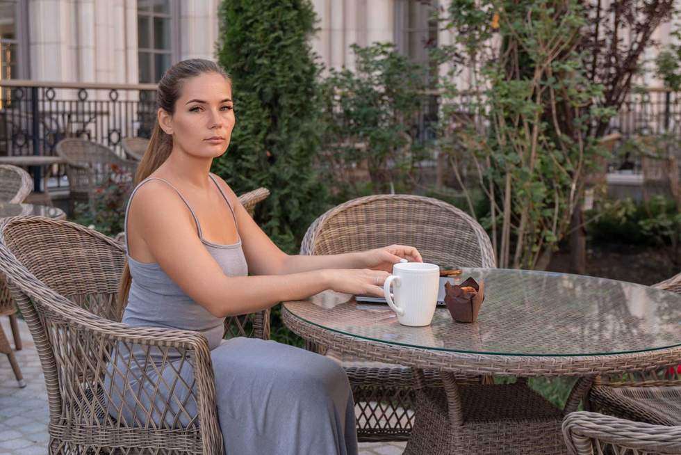 A woman sits at an outdoor cafe table with a cup of coffee and pastry. She is looking directly at the camera.