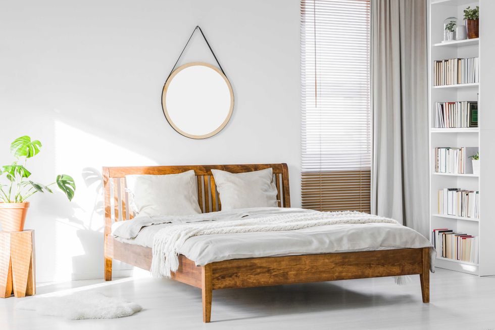 A wooden rustic bed frame and a home library bookcase in a natural, sunlit hotel room interior with white walls