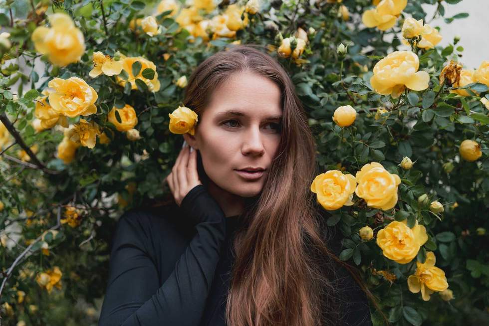 A young woman with long brown hair stands surrounded by blooming yellow roses in a garden.