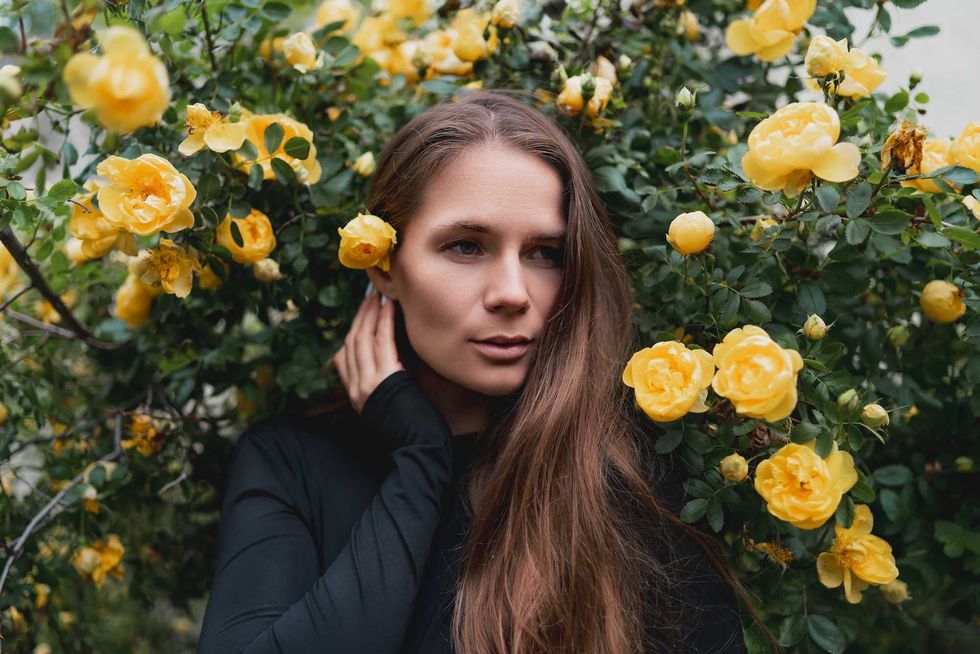 A young woman with long brown hair stands surrounded by blooming yellow roses in a garden.