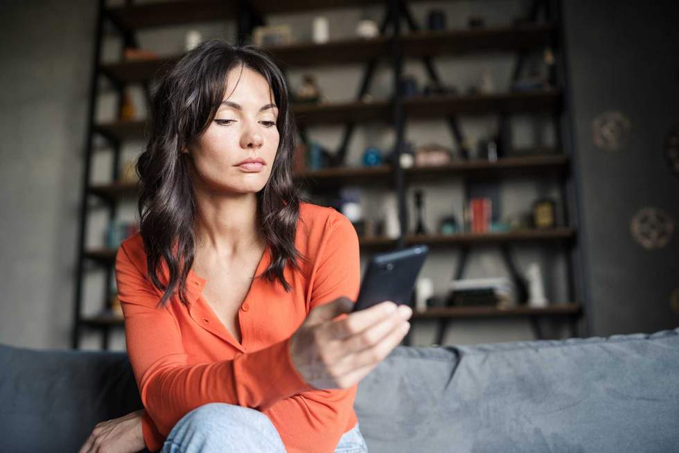 Attractive brunette woman sitting on sofa at home and thinking while typing or reading sms on cell phone. Beautiful female text messaging on mobile phone