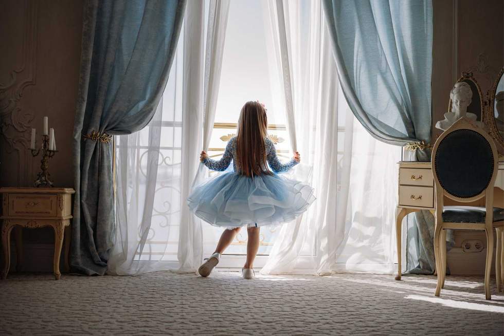 Back view of little girl in blue dance dress standing by the window, opening curtains in luxury apartment bedroom and looking out at the sea. Image with selective focus