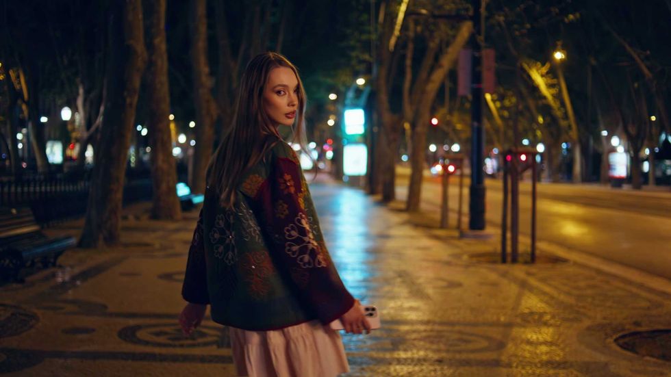 Back view walking girl turning to camera late evening outdoors. Confident young woman going on night street pavement illuminated streetlights. Gorgeous relaxed model strolling on urban neighbourhood