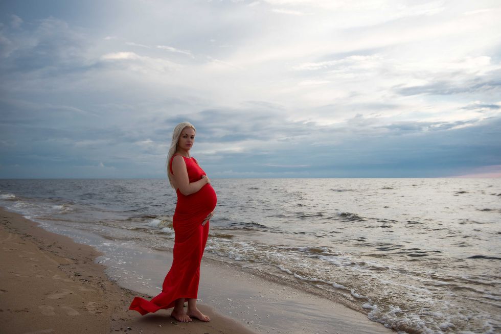 Beautiful pregnant woman in red dress standing at the beach during sunset