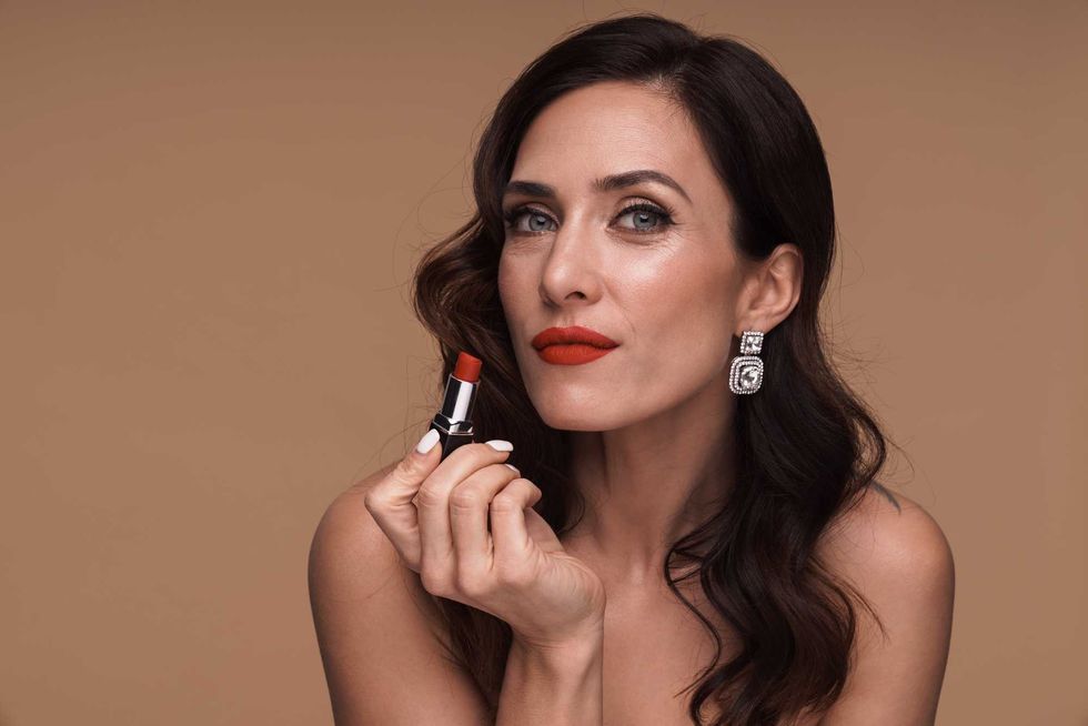 Beauty portrait of gorgeous woman applying red lipstick isolated over beige background