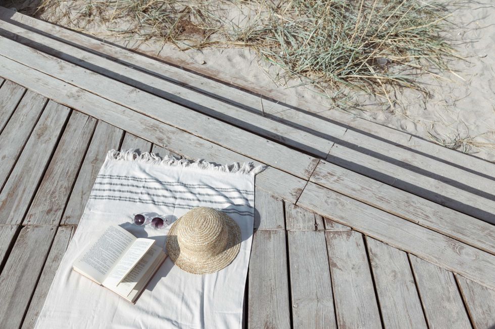 Boho eco styled beach essentials on sand by sea. Straw hat, glasses, book and beach towel on wooden backround, top view.